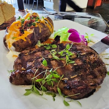 Steak with microgreens on a plate with a loaded baked potato, next to a wood plate of cornbread