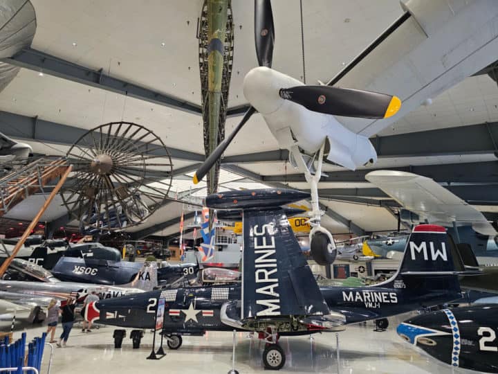 Blue and White Marines fighter jet with MW on the tail sitting on the museum floor, white airplane with propellers and other airplanes hanging from the ceiling 