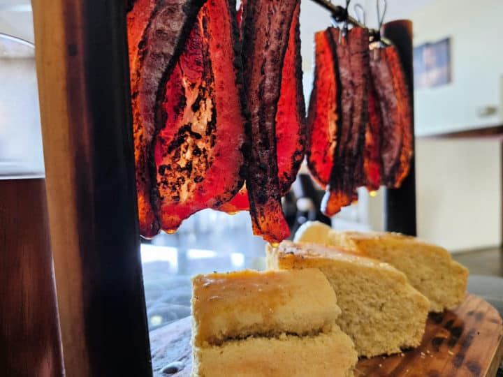 Bacon hanging from a wire dropping onto stacks of cornbread on a wooden board. 