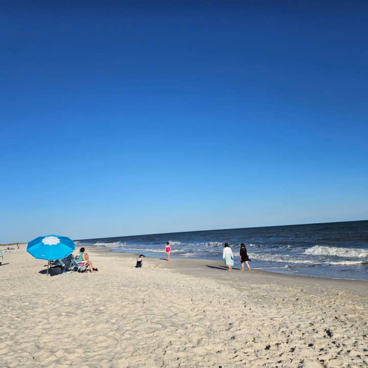 white sand beach with people walking down the beach near a blue umbrella and waves coming in on shore