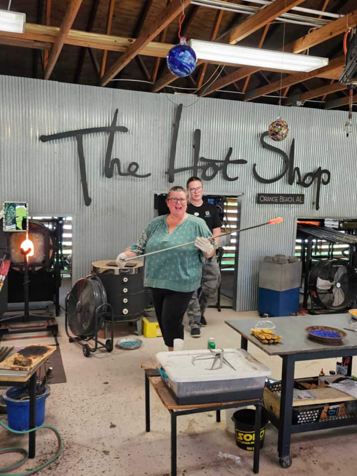 Tammilee holding a punty with hot glass on the tip next to a glass artist and The Hot Shop sign