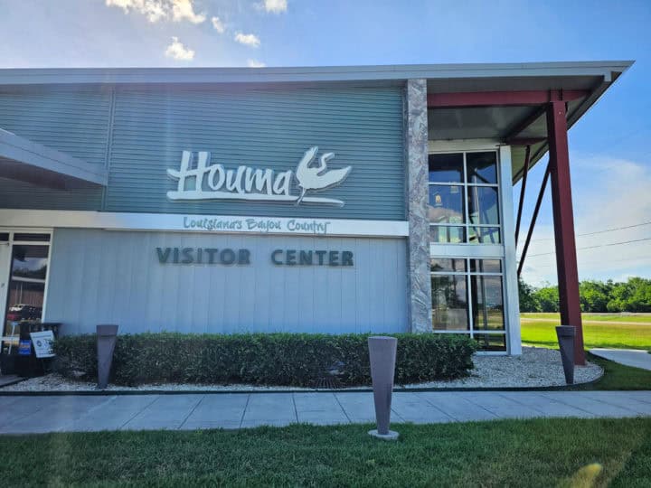 Houma Visitor Center on the side of a building with large glass windows and green bushes below the sign. 