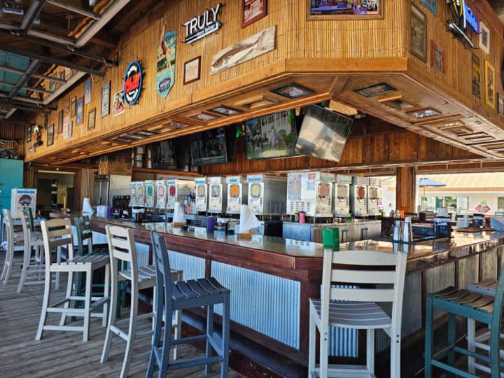 Tall bar chairs running in a line next to a bar with wood ceiling, and multiple frozen drink dispensers. 