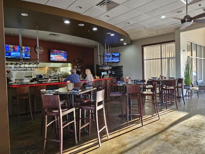 Indoor bar with TVs over it with tall chairs at the bar and multiple tall tables with tall chair nearby