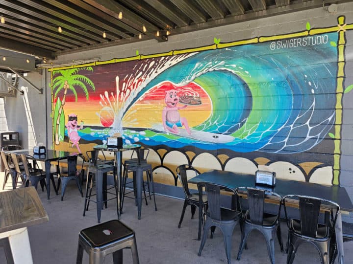 outdoor tables and chairs near a colorful mural with a surfing pig, large wave, and palm trees