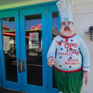 Chef statue with a chef hat with papa printed on it, chef coat that says Papa's pizza Pensacola Beach Boardwalk, next to a blue glass door entrance