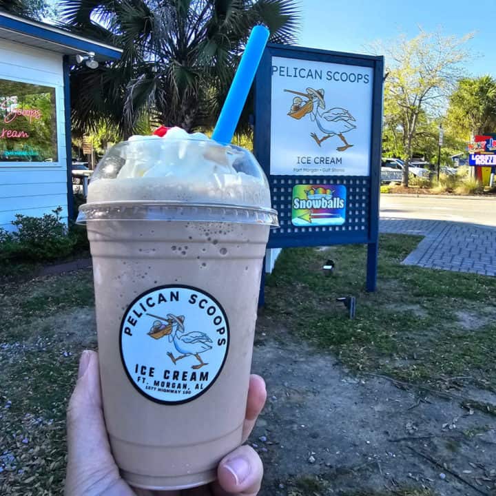 Hand holding a milkshake with a sticker showing Pelican Scoops Ice cream on the cup, next to a Pelican Scoops Ice Cream and Snowballs sign near a building. Tacky Jacks sign lit up in the background. 