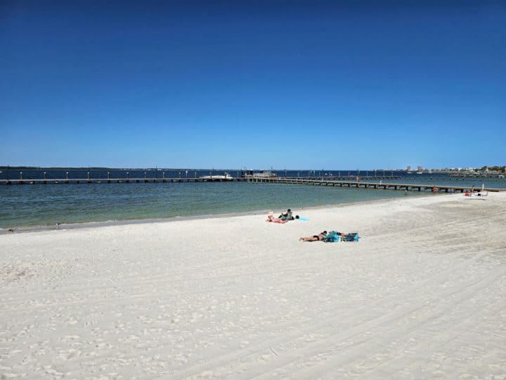 white sand beach with people sitting on it near the blue water with a pier in the background