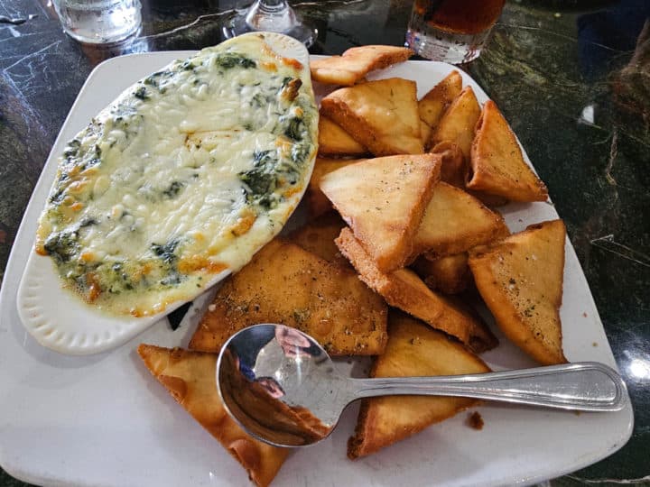 Spinach artichoke dip in an oval serving dish on a white square plate with large pita chips and a spoon