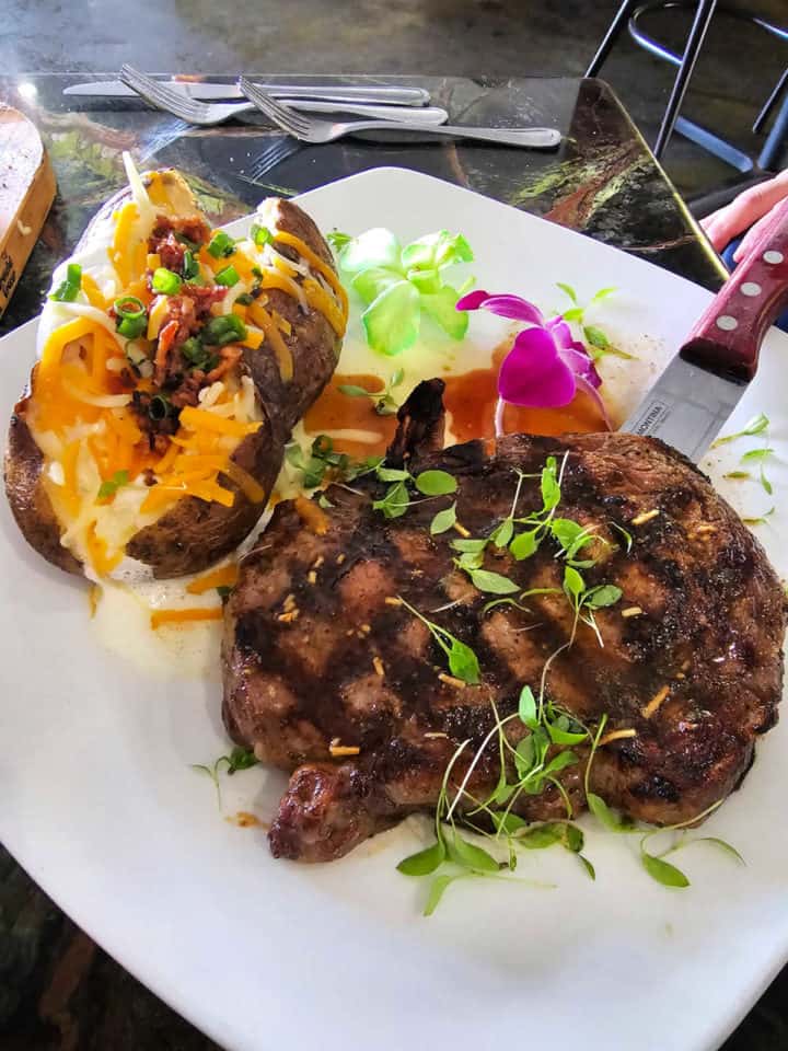 Steak and loaded baked potato on a square plate with a knife and purple orchid