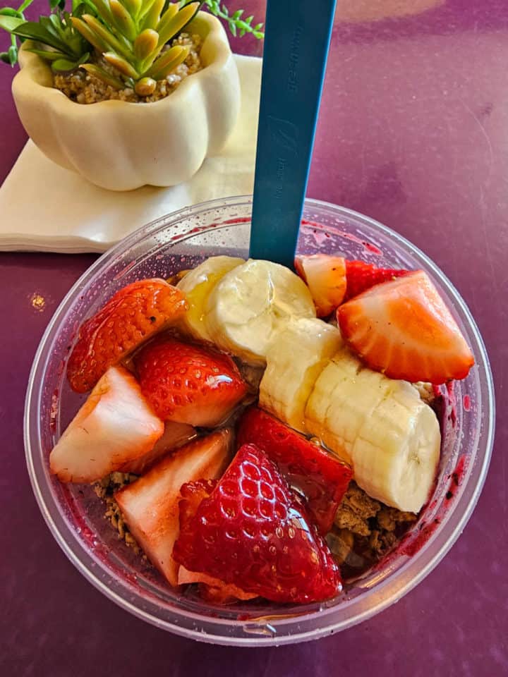 strawberry banana acai bowl in a plastic bowl with a spoon next to a succulent plant on a purple table