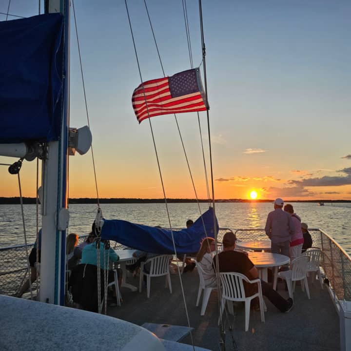 sunset over the water from a sailboat with American flag flying and people sitting on white chairs. 