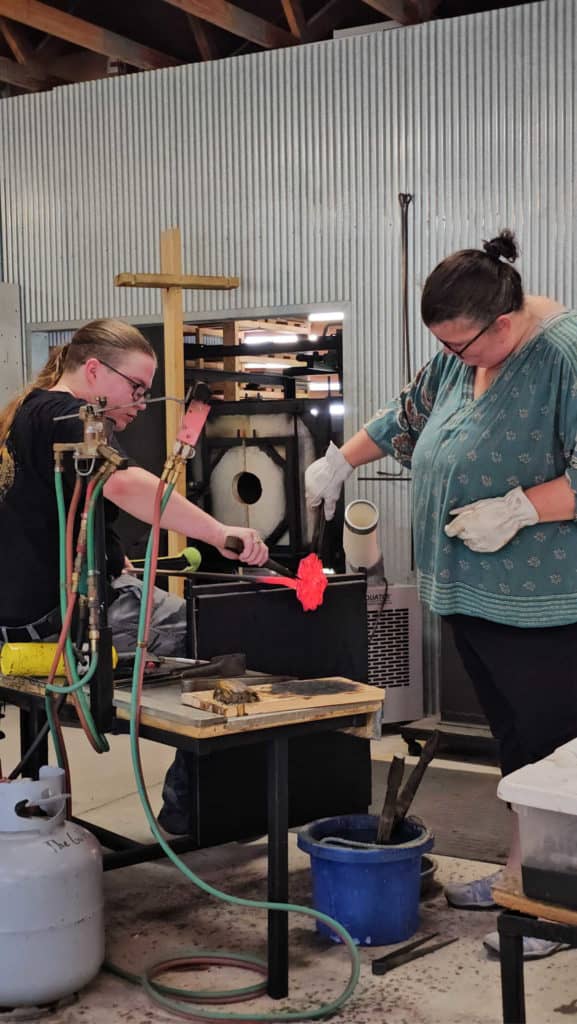 Tammilee pulling hot glass with large tweezers next to a glass artist and equipment