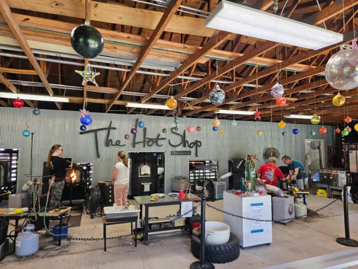 The Hot Shop Orange Beach sign on the wall near a furnace, four people working on glass, glass bulbs hanging from the ceiling, and glass blowing equipment
