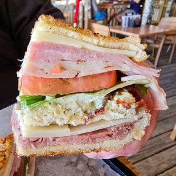 Large sandwich with ham, turkey, roast beef,lettuce, tomato, and cheese layered in pieces of bread being held above the table.