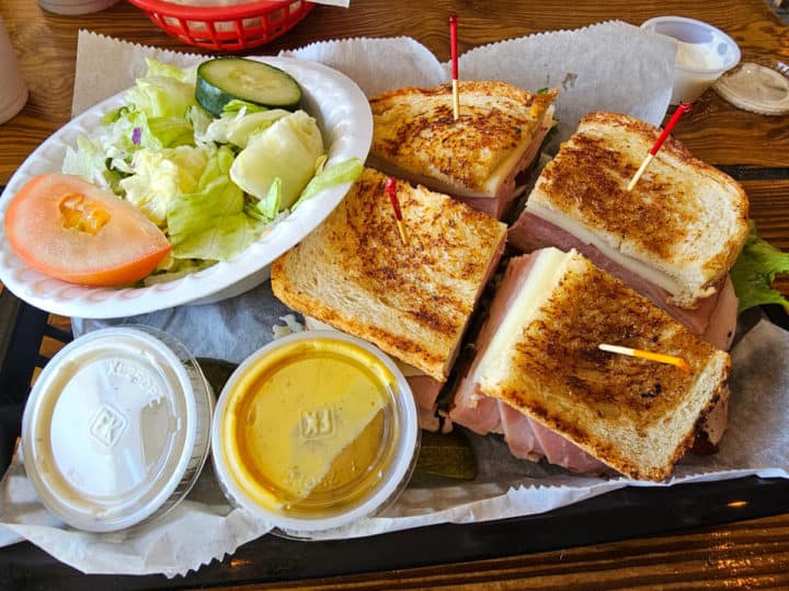 Large sandwich cut into quarters next to a side salad and two plastic containers of dressing in a paper lined basket