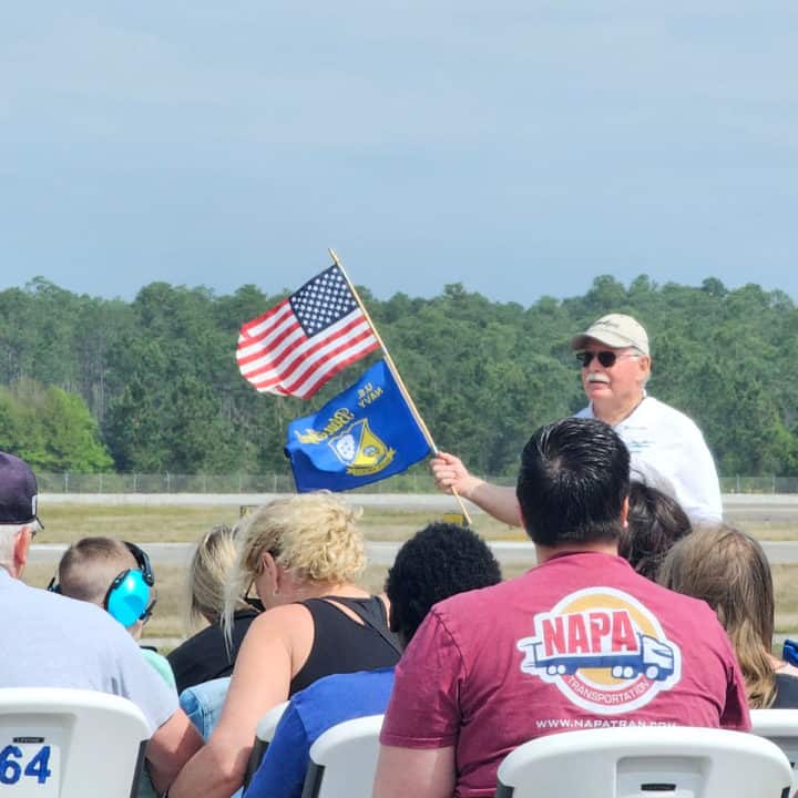 Older gentleman in white shirt holding a Blue Angels flag and American Flag in front of people sitting in chairs