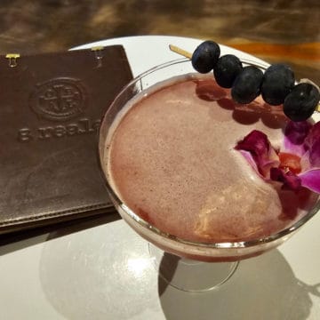 8 Reale leather menu next to a coupe glass with pink drink garnished with blueberries and an orchid.