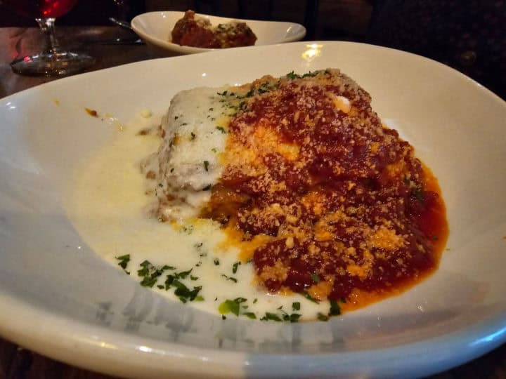 lasagna covered in red and white sauce on a white plate with meatballs in a bowl in the background
