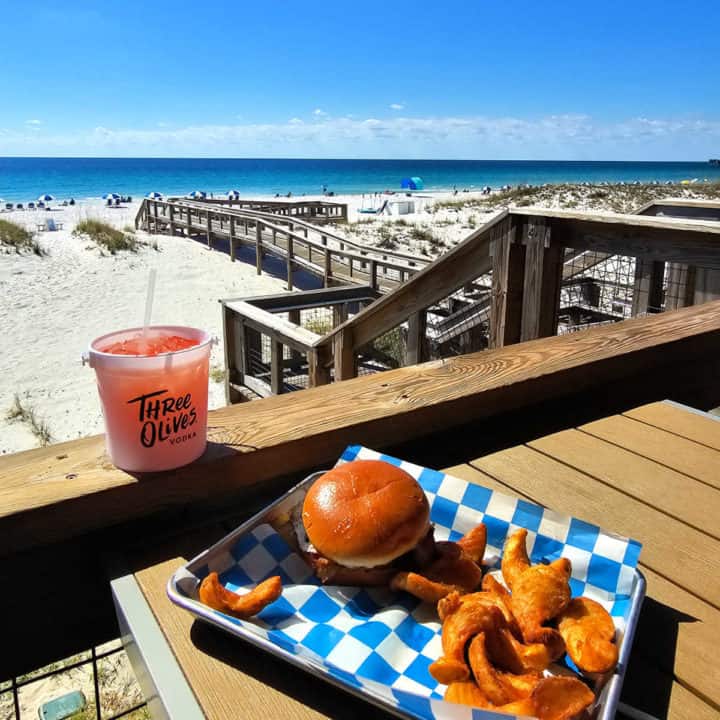 burger and fries on a platter next to a bucket drink looking out to the Gulf and white sand beaches. 