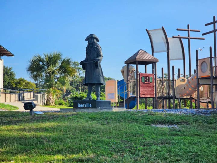 Iberville statue in front of kids playground equipment and a grassy area 