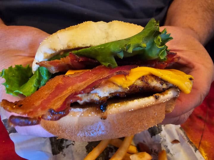 Grilled chicken sandwich with bacon, lettuce, and cheese being held above a table