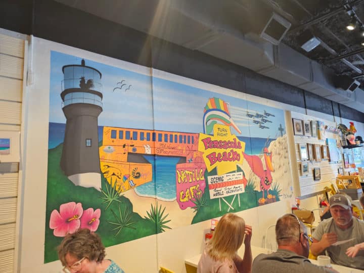 Pensacola Beach mural with historic sign, lighthouse, and flowers on the wall above tables