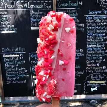 Pink popsicle with white and red heart sprinkles held in front of a menu board.