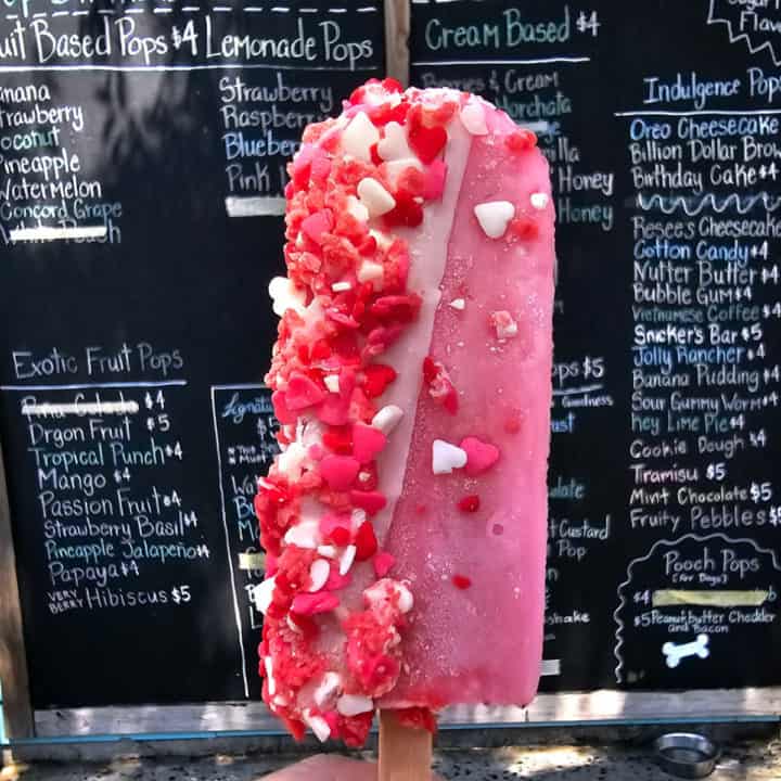 Pink popsicle with white and red heart sprinkles held in front of a menu board. 