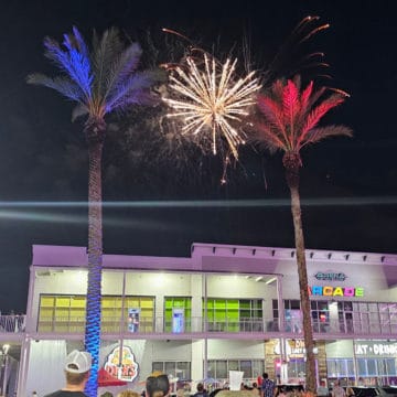 Palm trees lit up red and blue with white fireworks in the middle above stores at The Wharf