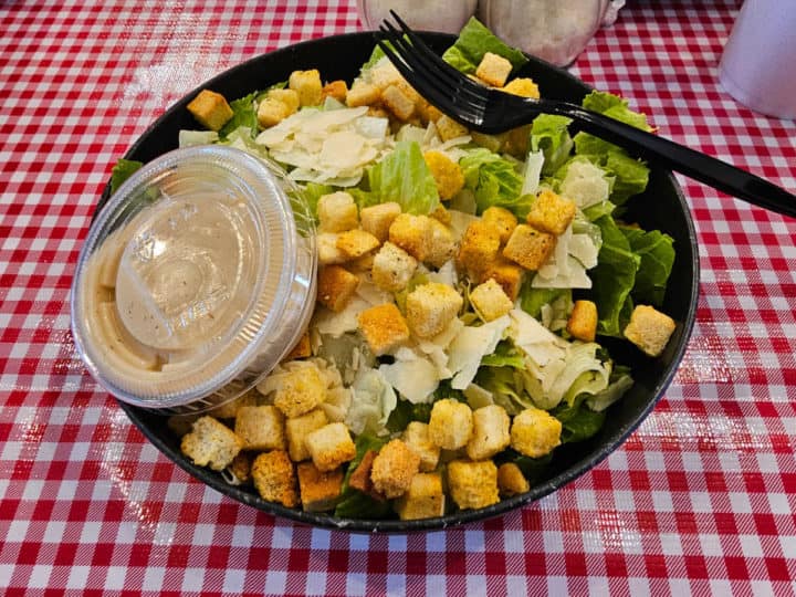 Caesar Salad in a black bowl with dressing on the side on a checkered tablecloth