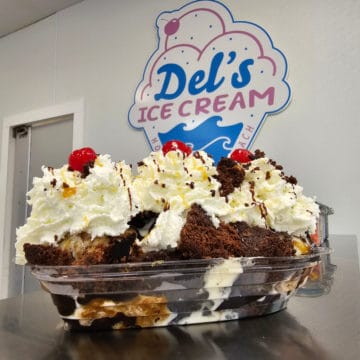 Brownie sundae with whipped cream and cherries in front of a Del's Ice Cream sign in the shape of an ice cream cone.