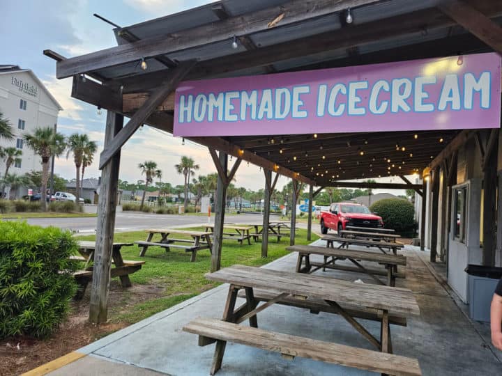 Homemade Ice Cream sign over a row of picnic tables under a shade cover with palm trees in the background