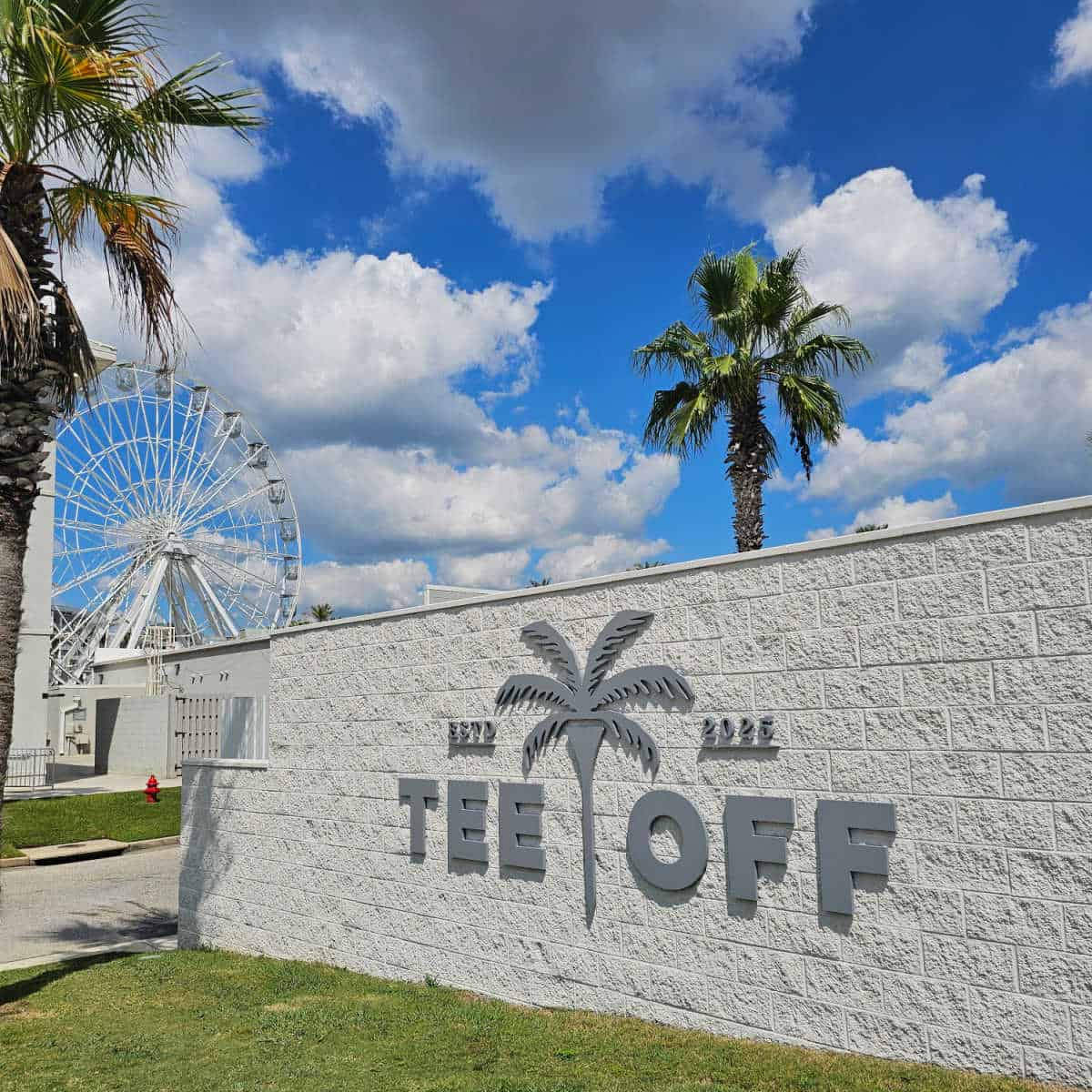 Tee off sign with palm tree logo on a brick wall with palm trees and Wharf Ferris Wheel behind it
