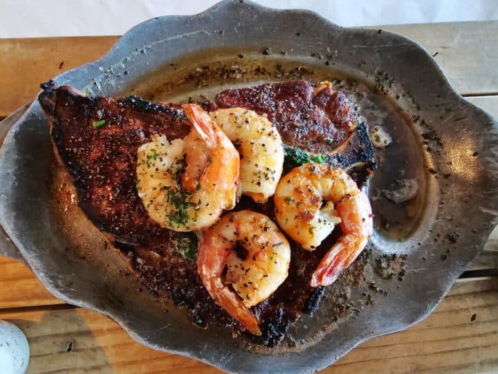 porterhouse steak with four Gulf shrimp on top sitting in a metal dish on a wooden board