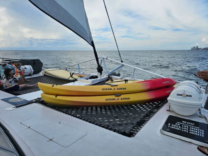 Kayaks on the front of the sail wild hearts sailboat with the sails up. People sitting near the kayaks