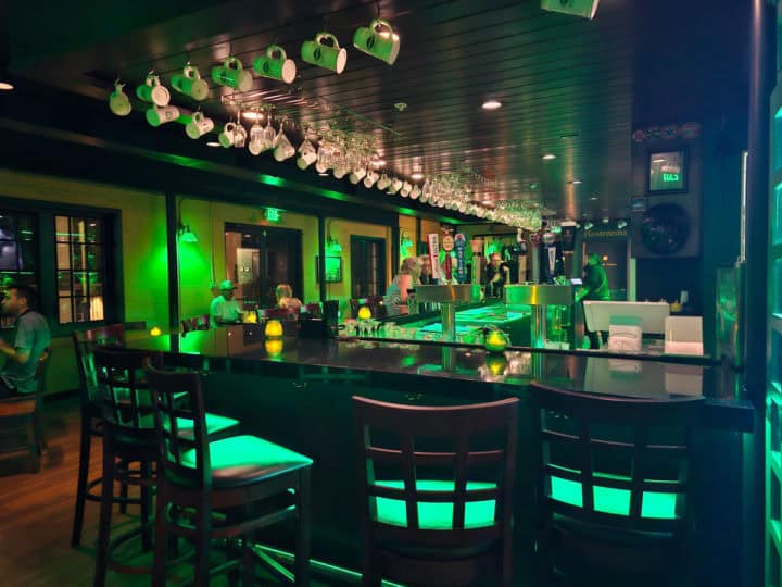 Long indoor bar with people sitting in tall chairs, beer mugs hanging from the ceiling, green lights in the pub