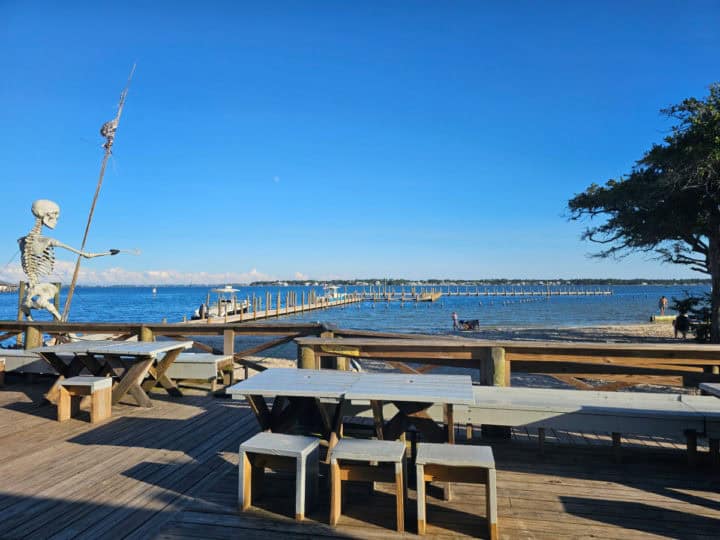 Outdoor tables and chairs on a deck looking out to water and a boat dock. Large skeleton next to the deck