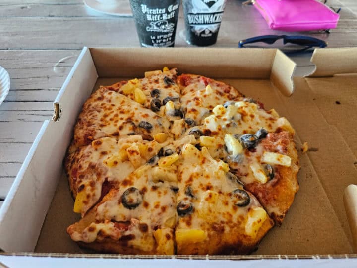 cheese and pineapple pizza in a cardboard box with bushwackers in the background. 