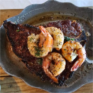 Gulf Shrimp on top of a porterhouse steak in a metal dish on a wooden board.