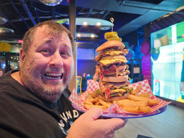 John holding a gigantic foot tall Big Kahuna Burger with a large television in the background