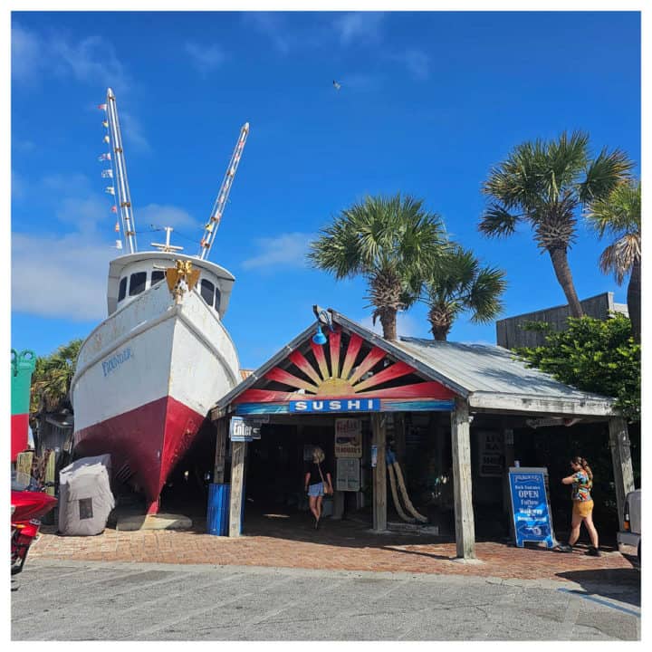 Replica boat with Flounders on it next to the restaurant entrance with a sushi sign, people walking to the entrance. 