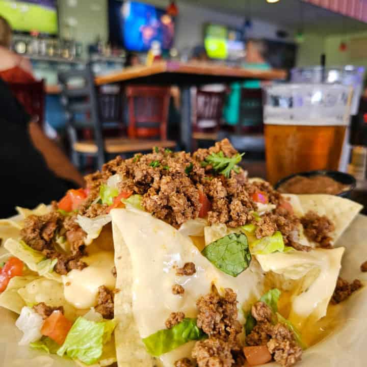 Plate of nachos with a glass of beer behind it on a table with TVs playing football games in the background