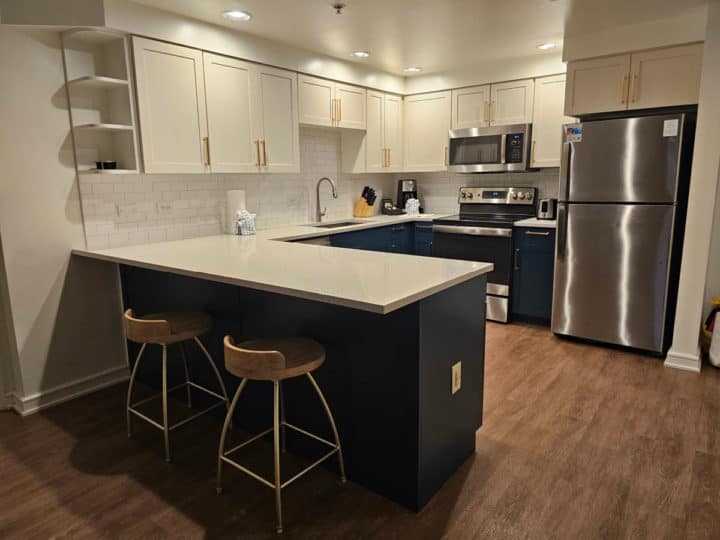 Kitchen with white cabinets on top, blue lower cabinets, two stools under the counter, and stainless steel appliances