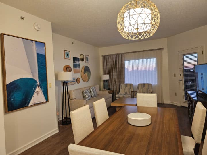 Dining room table with white chairs, coastal art on the wall, couch and chairs in front of a flat screen television. 