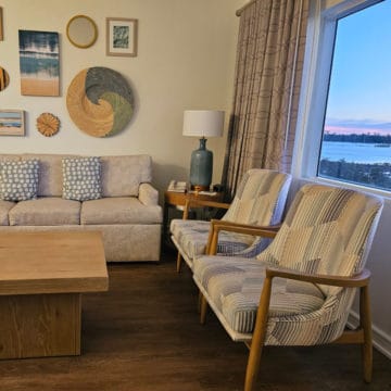 Two chairs next to a window looking out at the water, couch along the wall with coastal art above it