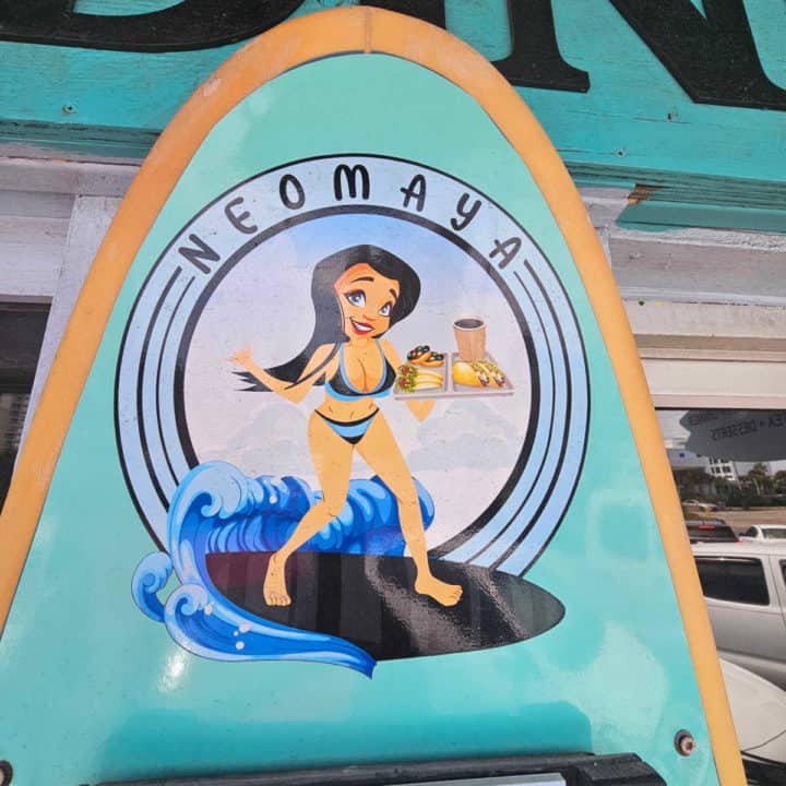 Neomaya logo with a brown haired woman surfing holding a platter of food and coffee printed on a surfboard