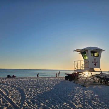 Lifegaurd stand lit up by the sunset with people playing on the beach by the water