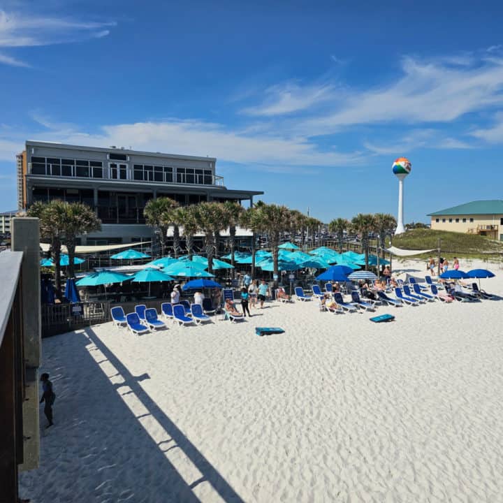 building with turquoise umbrellas in front of the restaurant, beach chairs on the beach, and Pensacola Beach water tower in the distance. 