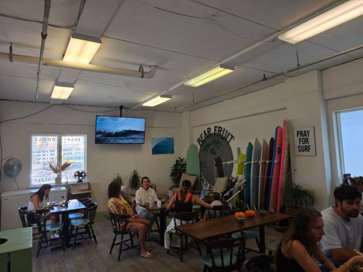 indoor seating with people at tables and chairs next to a rack of surfboards and a Bear Fruit mural on the wall